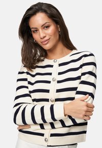 Gilet rayé blanc et marine avec encolure ronde, boutons proéminents, texture côtelée et poignets ajustés.