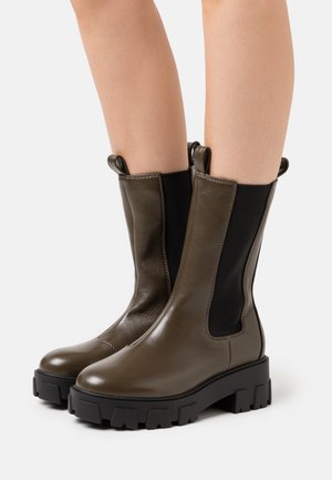 Bottines en cuir vert olive avec des panneaux latéraux élastiques noirs et des semelles en caoutchouc noir robustes. La semelle épaisse présente un motif de crampons.