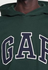 Donkergroene hoodie met grote, geborduurde 'GAP' letters in marineblauw met witte omlijning. Zachte katoen stof, casual ontwerp, en capuchon met trekkoord.