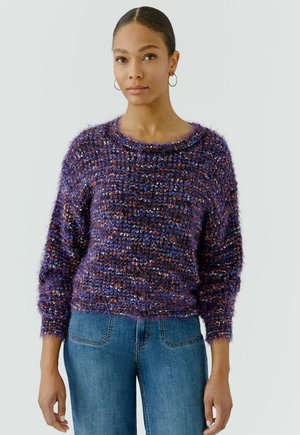 Vrouw met haar in een knot, die gouden oorringen draagt, een pluizige multicolor paarse trui en blauwe spijkerbroek, staand tegen een effen achtergrond.