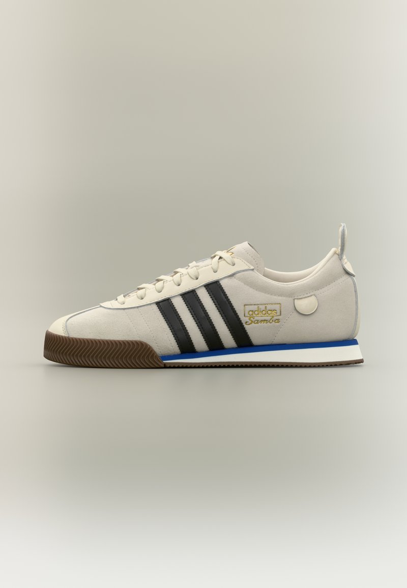 Väriltään melkein valkoinen Adidas Samba -lenkkikenkä, jossa on mustat raidat, kumipohja, sininen välipohjan raita ja kultaiset logotiedot sivulla.