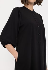 Černé dlouhé šaty s balónovými rukávy, knoflíkovou légou a bočními kapsami. Hladká textura; minimalistický design.