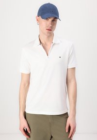 Witte polo met een ritssluiting bij de kraag, voorzien van een gedetailleerd logo op de borst, gecombineerd met een blauwe baseballcap en groene shorts.