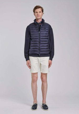 Uomo che indossa un gilet imbottito blu navy, una camicia nera a maniche lunghe, pantaloncini bianchi arrotolati, scarpe da barca blu navy e una sciarpa a motivi al collo, in piedi contro uno sfondo semplice.