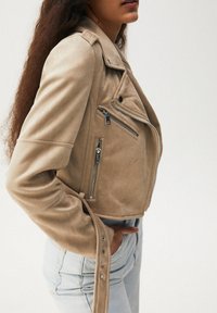 Veste de motard en daim beige avec fermeture éclair asymétrique, poches latérales et détails à boutons-pression sur les manches ; coupe courte mettant en valeur une silhouette ajustée.
