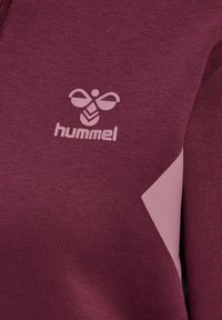 Vínová mikina s kapucí, která má světle růžový trojúhelníkový akcent a embosované logo. Vyrobena z měkké látky, s designem límce na polovinu zipu.