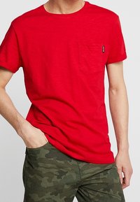 Rød bomulds T-shirt med rund halsudskæring og korte ærmer, der har en lomme på venstre bryst og et lille mærke. Bæres med camouflagebukser.