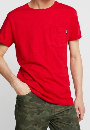 Röd bomulls t-shirt med rund krage och korta ärmar, med en ficka på vänster bröst och en liten märkesetikett. Bärs tillsammans med kamouflagebyxor.