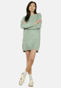 Svetlozelené svetrové šaty z vlny s dlhými rukávmi, bočnými rozparkami a voľným strihom, doplnené o krémové tenisky a šedé ponožky.
