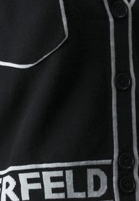 Garbamento nero con chiusura frontale a bottoni e due bottoni, dettagli bianchi a contrasto lungo il colletto e la lista dei bottoni, e un logo.