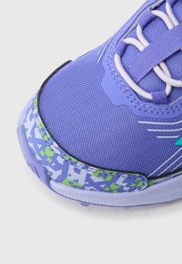 Μωβ αθλητικό παπούτσι με υφασμάτινο επάνω μέρος με υφή, διαθέτει πράσινο και μπλε σχέδιο στη σόλα και αντίθετα ανοιχτόμπλε στοιχεία.