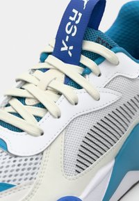 Sapatilha desportiva em branco e azul claro, com painéis de malha, detalhes texturizados e uma alça azul com a marca "RS-X" em destaque.