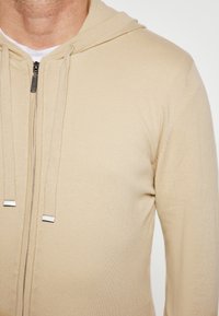 Tan zip-up hoodie met een zachte textuur, voorzien van een verstelbare capuchon en zilveren hardware accenten. Het is ontworpen om strak om het lichaam te passen.