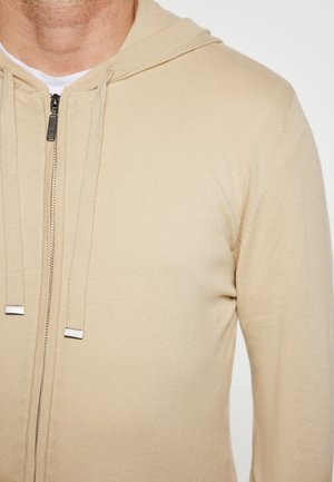 Tan zip-up hoodie met een zachte textuur, voorzien van een verstelbare capuchon en zilveren hardware accenten. Het is ontworpen om strak om het lichaam te passen.