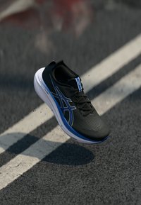 Chaussure de sport noire avec une tige texturée, accents bleus et une semelle blanche, présentée en plein vol au-dessus d'une surface grise avec des marquages routiers.