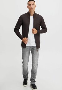 Brun ribbet zip-up cardigan med høj krave, bæret over en hvid t-shirt, parret med lysegrå jeans og sorte sneakers.