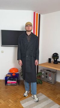 Lungo cappotto doppiopetto nero con grandi revers, abbinato a una camicia a righe, jeans blu e scarpe da ginnastica con motivo a fantasia. Tavolo arancione sullo sfondo.