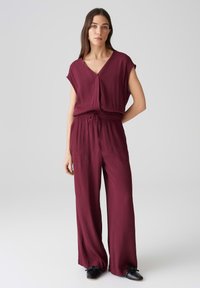 Bordeauxfarbener Overall mit kurzen Ärmeln, V-Ausschnitt, elastischem Taillenbund und weitem Bein. Glatte Textur mit Vordertaschen und Kordelzugdetails.