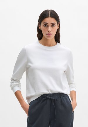 Femme aux cheveux foncés portant une chemise blanche à manches longues et un pantalon bleu marine à cordon, debout les mains dans les poches sur un fond blanc.