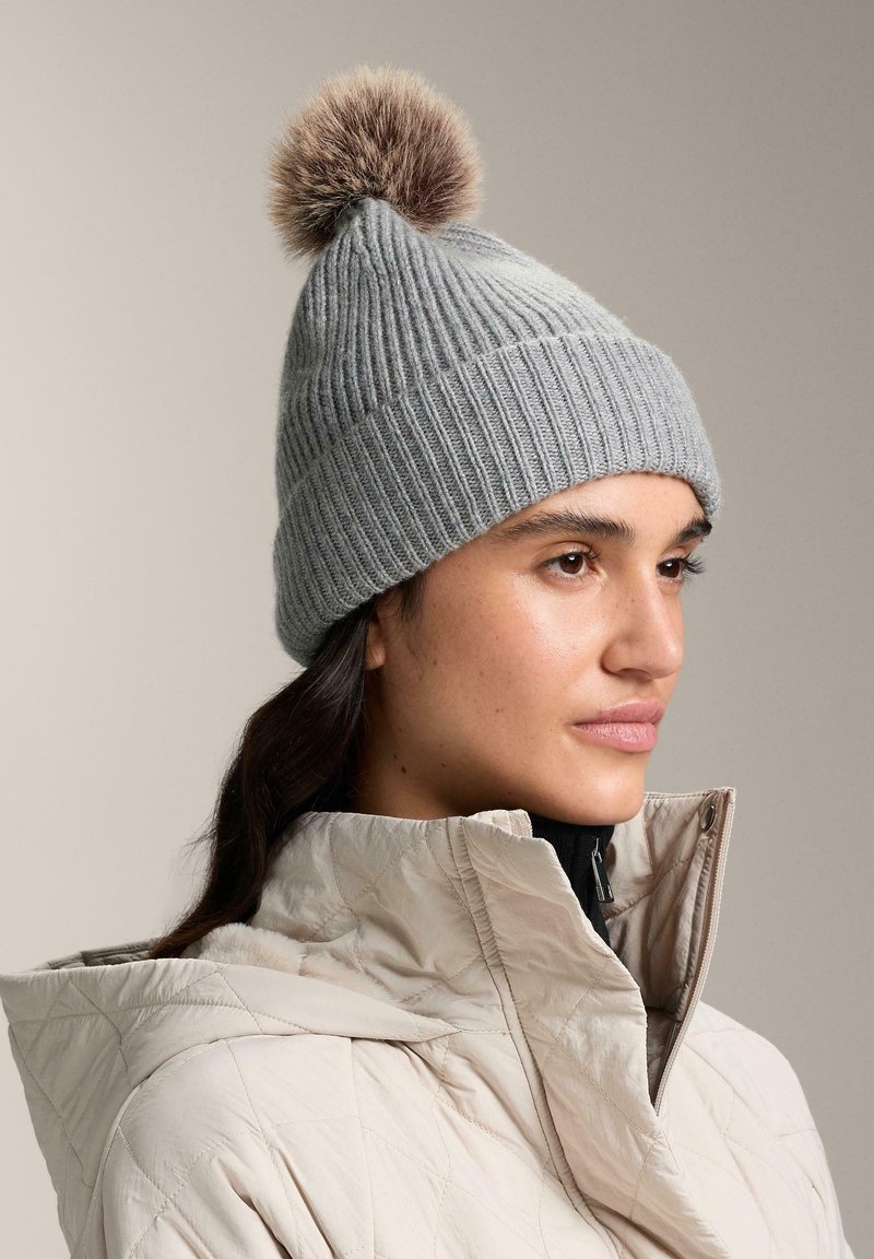 Bonnet gris tricoté avec une texture côtelée et un pompon en fourrure sur le dessus, porté avec une veste matelassée beige dotée d'un col montant.