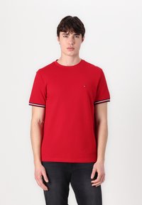 Červené bavlněné tričko s kulatým výstřihem a krátkými rukávy se pruhovanými detaily. Model nosí tmavé džíny. Jednoduchý design se malým logem.