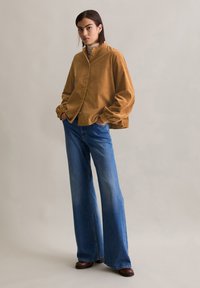 Chemise boutonnée en velours côtelé marron avec une coupe décontractée et des manches longues froncées, portée avec un jean en denim bleu à jambes larges et des bottes noires.
