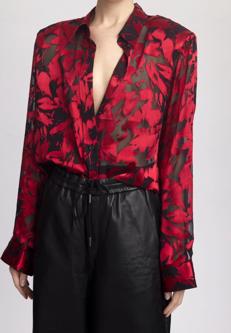Rode en zwarte bloempatron blouse van doorschijnende stof met een diepe V-hals en knoopsluiting, gecombineerd met zwarte leren high-waisted broeken.