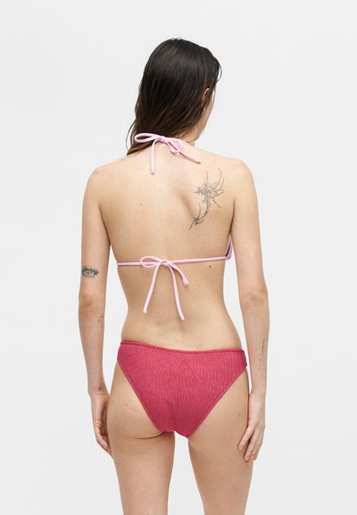 Γυναίκα με μακριά σκούρα μαλλιά που φορεί ροζ μπικίνι τοπ και υφασμάτινο κόκκινο κάτω μέρος μπικίνι, με τατουάζ στην πλάτη και αριστερό χέρι, στραμμένη προς τα πίσω.