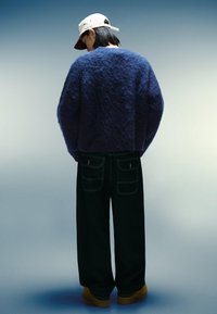 Oversized marinblått fluffigt tröja, svarta vida jeans och beige skor. En vit och röd keps fulländar outfiten. Bakifrån mot en gradientbakgrund.