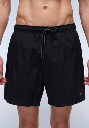 Mandlig overkrop iført sorte badeshorts med snøre og et lille norsk flagemblem på venstre ben, mod en neutral baggrund.