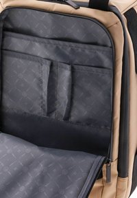 Interno di uno zaino beige con rivestimento grigio, caratterizzato da diverse tasche e un sottile motivo di cerchi e frecce. Compartimenti con zip visibili.