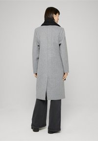 Sivi vuneni kaput s dugim, ravnim krojem, crnim ovratnikom i prorezom na stražnjoj sredini, u paru s širokim crnim hlačama.