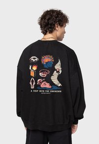 Sudadera negra de gran tamaño con gráficos multicolores en la parte posterior, que incluyen diseños abstractos, texto y varios íconos simbólicos.