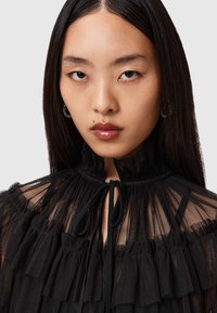 Blusa nera trasparente con ruches, colletto alto e dettaglio del collo annodato. Presenta una delicata texture e un design in tessuti a strati.