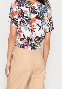 Short-sleeve blouse with a white background featuring a vibrant tropical leaf print in pink, orange, green, and black. Paired with tan pants.