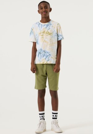 Garçon debout, portant une chemise à manches courtes à motif abstrait, un short vert olive, des baskets montantes blanches et des chaussettes blanches avec des lettres noires.