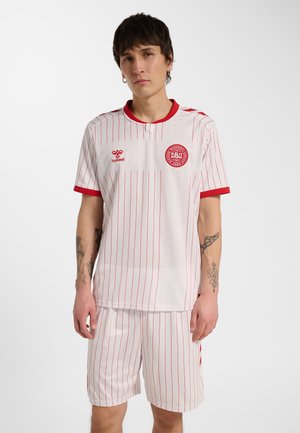Ung person iført hvid fodboldtrøje og shorts med tynde lodrette røde striber og rød kant, stående mod en ensfarvet baggrund.