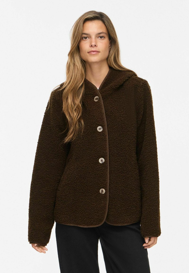 Femme portant une veste en polaire teddy marron foncé boutonnée, manches longues et coupe ample, debout devant un fond clair uni.