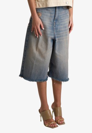 Persona che indossa pantaloncini di jeans sbiaditi a vita alta, lunghi fino al ginocchio con orli sfrangiati e sandali beige con tacco alto e cinturini.