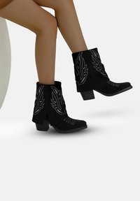 Botas de tobillo de ante negro con bordado plateado en forma de alas, un tacón apilado y un diseño de punta afilada. Textura suave con detalles minuciosos.
