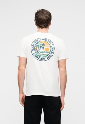 Άνδρας γυρισμένος με την πλάτη φοράει λευκό μπλουζάκι με στρογγυλό πολύχρωμο γραφικό παραλίας Santa Cruz που περιλαμβάνει έναν φοίνικα, κυματισμό του ωκεανού και τον ήλιο που δύει.