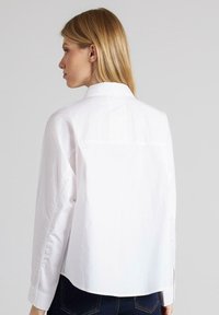 Camisa blanca de botones con mangas largas, cuello y corte recto. Presenta una textura suave y un diseño de canesú en la parte posterior.