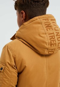 Braune Kapuzenjacke mit matter Oberfläche. Sie verfügt über sichtbare Nähte, eine Seitentasche mit einem Druckknopf und Textdetails auf der Kapuze.