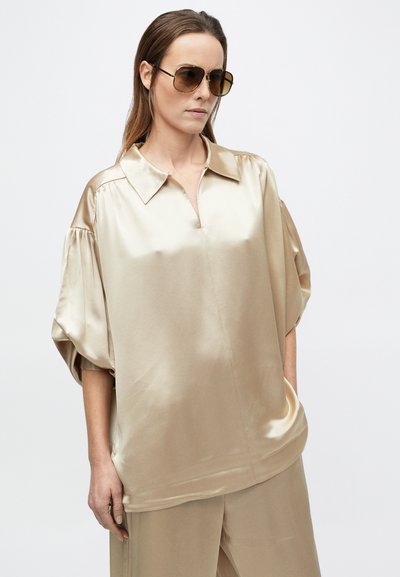 Nő túlméretezett, fényes bézs színű szatén blúzban, félig hosszú ujjakkal és hozzávaló, bő szárú nadrágban, pilóta napszemüveggel kiegészítve.