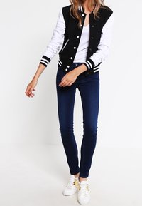 Chaqueta varsity en blanco y negro con puños a rayas, combinada con jeans oscuros ajustados y zapatillas blancas, con una tela texturizada.