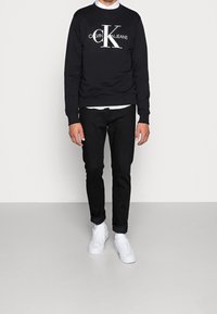 Černá mikina s bílým logem "CALVIN KLEIN JEANS", kombinovaná s tmavými džíny a bílými teniskami. Jednoduchý design s kulatým výstřihem.