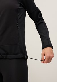 Giacca sportiva nera in tessuto testurizzato, con una tasca laterale con zip e cordino regolabile all'orlo. Maniche lisce.