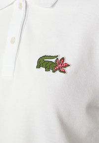 Camisa polo blanca con un cocodrilo verde y una flor roja bordados en el lado izquierdo del pecho. El material tiene un acabado texturizado.