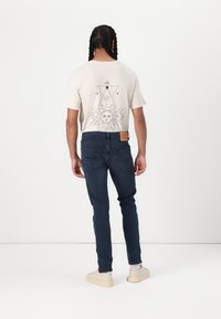Beige rövidujjú póló nap grafikai mintával, sötétkék farmernadrággal párosítva. A modellen krémszínű, texturált talpú sportcipő van.