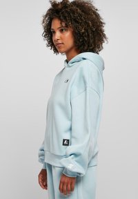 Light blue hoodie with a relaxed fit, featuring a front pocket, drawstring hood, and small logo on the chest. Soft fabric texture.